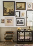 Entry hall with wood and leather chair, dark wood wall table with mirrored front, lamps, flowers and tray in front of light wall with various mirrors, paintings and photos.